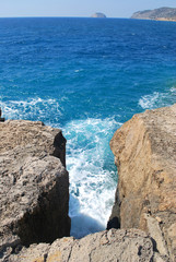 A crevice between large stones washed by sea waves with white foam against a blue sky, Cape Fourni, Rhodes Island, Greece