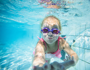 Fototapeta premium Wide angle underwater photo of a toddler girl swimming in a big swimming pool with goggles and a pink bikini