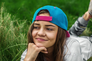 Beautiful white Caucasian girl in a baseball cap lies on a green grass on a summer day and looks away