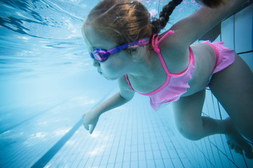 Wide angle underwater photo of a toddler girl swimming in a big swimming pool with goggles and a...