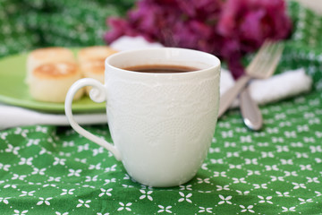 Black coffee in a white sophisticated cup, delicious dietary cheesecakes from home-made farmer cheese for breakfast, a bouquet of purple tulips on a green tablecloth.