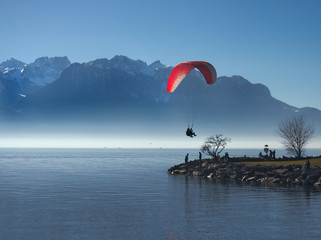 Paraglider tandem landing on little peninsula in lake with alpine mountains in the background.