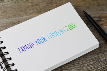 EXPAND YOUR COMFORT ZONE. hand lettering in notebook with pen on wooden background