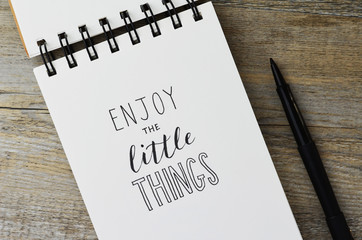 ENJOY THE LITTLE THINGS hand lettering in notebook with pen on wooden background