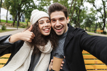 Fototapeta premium Happy young loving couple drinking tea or coffee outdoors in park talking with each other take a selfie by camera.