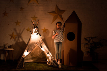 Kid standing between wigwam and cardboard rocket in dark room © LIGHTFIELD STUDIOS