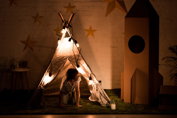 Kid standing on all fours in wigwam and looking at cardboard rocket in dark room © LIGHTFIELD STUDIOS