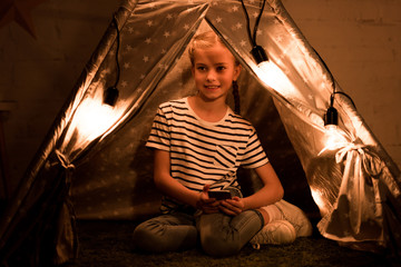 Happy kid with smartphone sitting in cozy wigwam at home © LIGHTFIELD STUDIOS