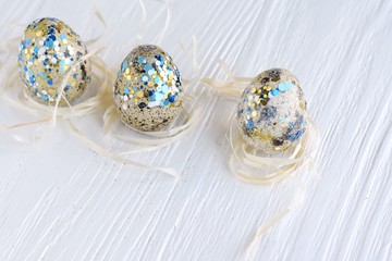 Tree spotted quail Easter egg with blue shiny creative decor and natural straw nest with selective focus on white wooden rustic background. Easter eggs and empty space for text