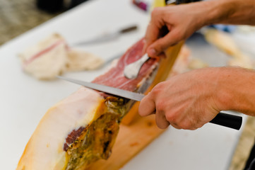Closeup on person hands slicing carving delicious dry-cured ham prosciutto, prepared delicatessen food culinary background.