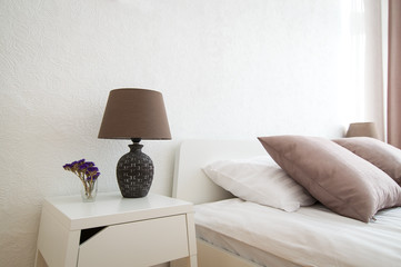 Interior design of bedroom. Nightstand and table lamp beside a bed with pillows. Matching white and coffee color shades