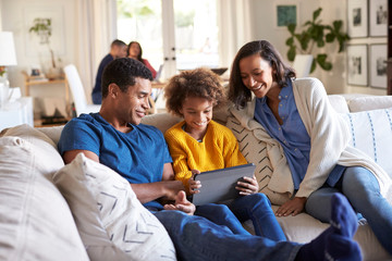 Young parents and their pre-teen daughter sitting on a sofa in the living room using tablet computer together, grandparents sitting at a table in the background