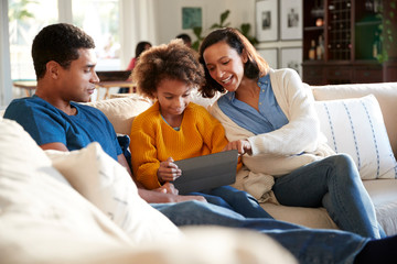 Young parents and their pre-teen daughter sitting on a sofa in the living room using a tablet computer together, selective focus