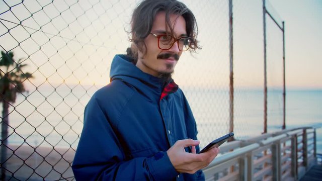 Young millennial hipster man with red glasses and long hair tail using mobile phone or smartphone application or app checking social network news feed to be up to date and being connected to internet