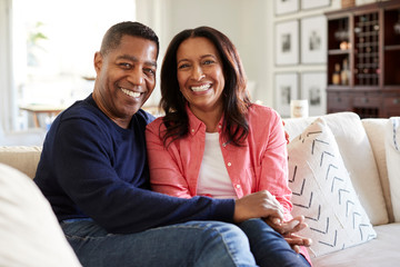 Middle aged mixed race couple sitting on the sofa in their living room looking to camera, front...