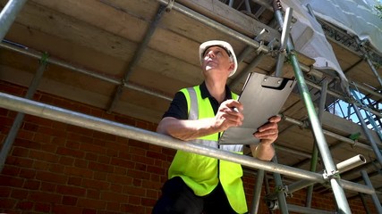 Slow motion panning shot of male builder foreman, worker or architect working on construction building site standing on scaffolding writing on clipboard 