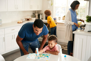 Dad watching his toddler son painting a picture sitting at a table in the kitchen, while mother and girl prepare food in the background