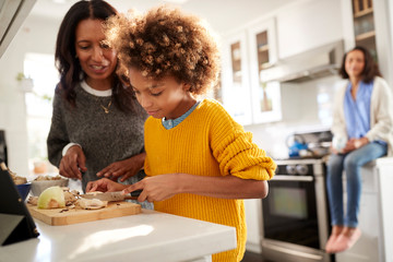 Grandmother helping her granddaughter prepare food in the kitchen, mother sitting in the...