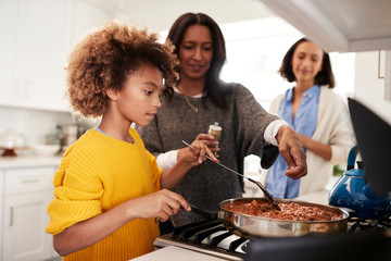 Pre-teen black girl standing at the hob in the kitchen preparing food with her grandmother and mother, close up, selective focus