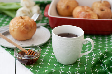 Baked apples in red, a white cup of tea or coffee, honey, a bunch of white tulips lie on a green tablecloth.