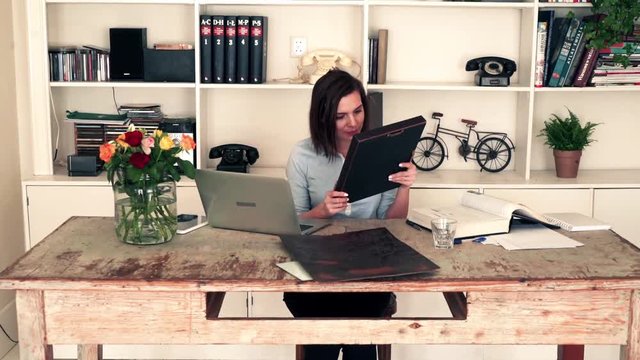 Young art curator looking at painting and checking something on laptop and book