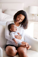 Young adult mother sitting in an armchair in her bedroom, holding her three month old baby son in...