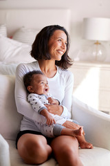 Young adult mother sitting in an armchair in her bedroom, holding her three month old baby son in...