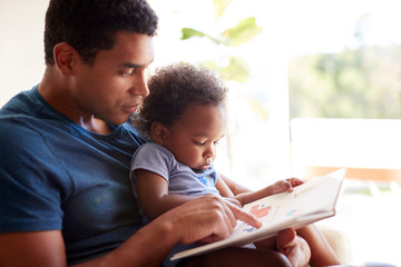 Close up of young adult black father reading a book with his two year old son, close up, side view,...