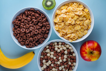 Cereals bowl of glazed, chocolate balls, rings, corn flakes and fresh ripe fruits for dry breakfast on blue background.