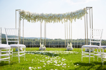 Stylish wedding arch for exit registration under the open sky. The lawn is covered with petals of white roses.