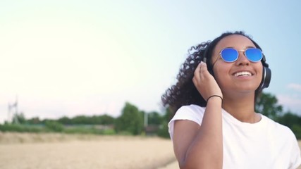 Slow motion follow shot video of beautiful mixed race African American girl teenager young woman in white T-shirt and blue sunglasses walking listening to music on wireless headphones 