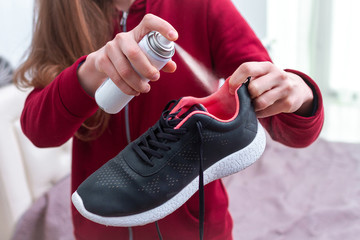 A young woman spraying deodorant on sweaty running shoes for eliminate unpleasant, bad smell. Shoe shine and care. Sport footwear needs in cleaning and odor removal. - obrazy, fototapety, plakaty