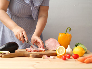 A woman cuts meat on a board, next to fresh vegetables lie: raw eggplant, carrots, lettuce, paprika, lemons, greens close-up on a light background