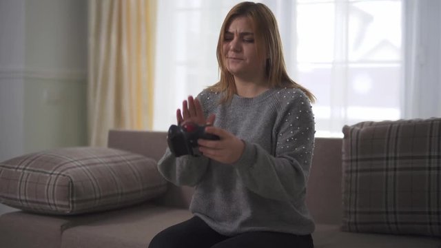 Plump girl playing video game sitting at sofa in home. Discontented chubby woman beats hand on broken joystick. Leisure of plus size lady at home indoors