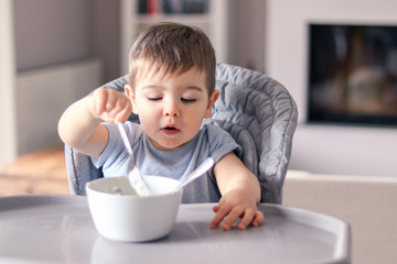 Cute little baby boy with funny smeared face concentrated on food eating with fork from white bowl at table in front of him sitting in high feeding chair. Attempt to eat by himself. Child nutrition