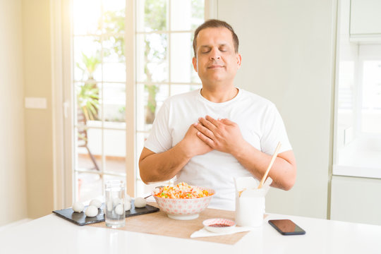 Middle age man eating asian food with chopsticks at home smiling with hands on chest with closed eyes and grateful gesture on face. Health concept.