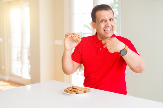 Middle age man eating chocolate chips cookies at home with surprise face pointing finger to himself