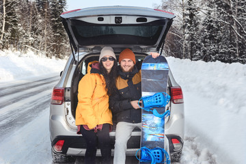 Smiling couple is sitting in the back trunk of suv car on roadside of winter road. Family trip to ski resort. Winter holidays adventure.  Man holding his snowboard. travelling lifestyle concept.