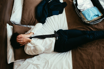 top view of exhausted businessman resting on bed with travel bag in hotel room