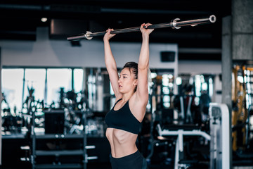 Young woman in sportswear doing shoulder press exercise with a weight bar at the gym.