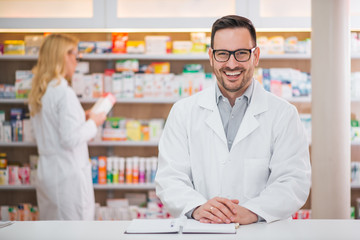 Portrait of a handsome pharmacist at the counter of a drugstore, female colleague working in the...