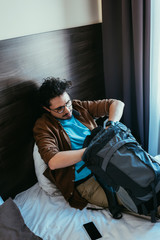 male tourist taking something from backpack while sitting on bed with smartphone in hotel