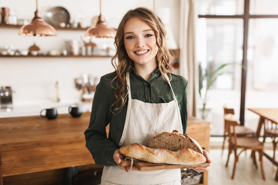 Young pretty smiling woman with wavy hair in white apron holding bread on board in hands happily looking in camera spending time in cozy kitchen