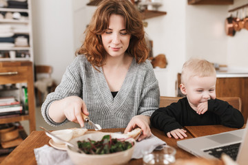 Young beautiful woman with red hair in sweater dreamily cooking with her little son near happily watching cartoons on laptop. Mom spending time with baby in kitchen at home