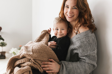 Young gorgeous mom with red hair in knitted sweater sitting on floor holding her little joyful son covering with blanket happily looking in camera together spending time at home