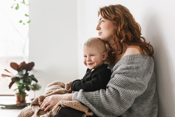 Young gorgeous mom with red hair in knitted sweater sitting on floor holding her little smiling son covering with blanket happily looking aside together spending time at home