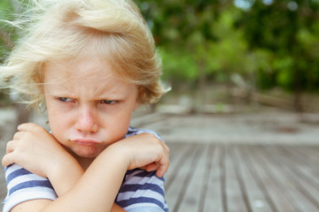 Face portrait of annoyed and unhappy caucasian kid with crossed arms. Upset and angry child concept for family relations, social problems issues and juvenile psychology.  Copy space outdoor background - obrazy, fototapety, plakaty