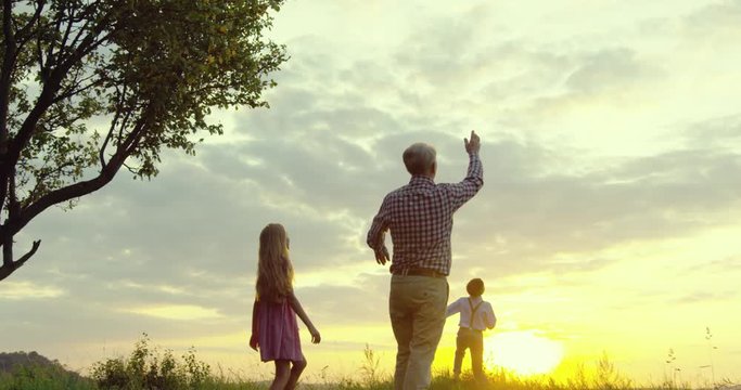 Happy and cheerful Caucasian senior grandfather with two kids - grandson and granddaughter - playing with plane toy model and launching it in the field on the picturesque sunset.