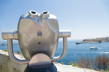 In the foreground a coin operated telescope in the background a view of the city of Kalkara, Malta