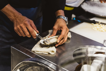 Chef shucking a fresh oyster by knife on stainless steel counter bar. 